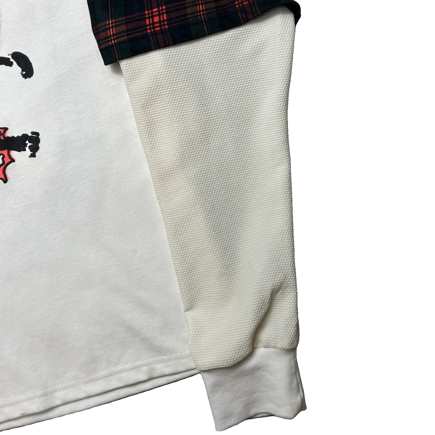A folded white T-shirt shown on a plain white background, with a black, red, and green plaid lining visible along one edge and a partial graphic print in black and red on the front.