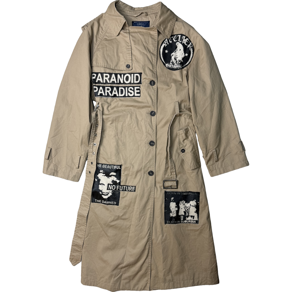 A beige trench coat with black buttons, featuring multiple black and white graphic patches including 'PARANOID PARADISE' and 'THE BEAUTIFUL NO FUTURE'.