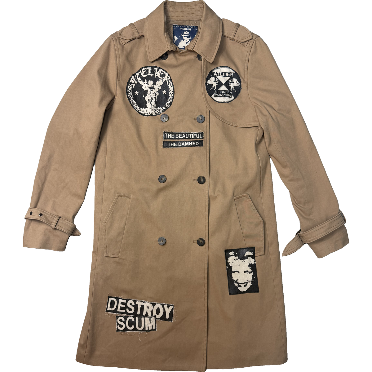 A tan double-breasted trench coat with multiple black and white patches, including 'THE BEATIFUL THE DAMNED', 'DESTROY SCUM', and 'KELLY'S', shown against a plain white background.