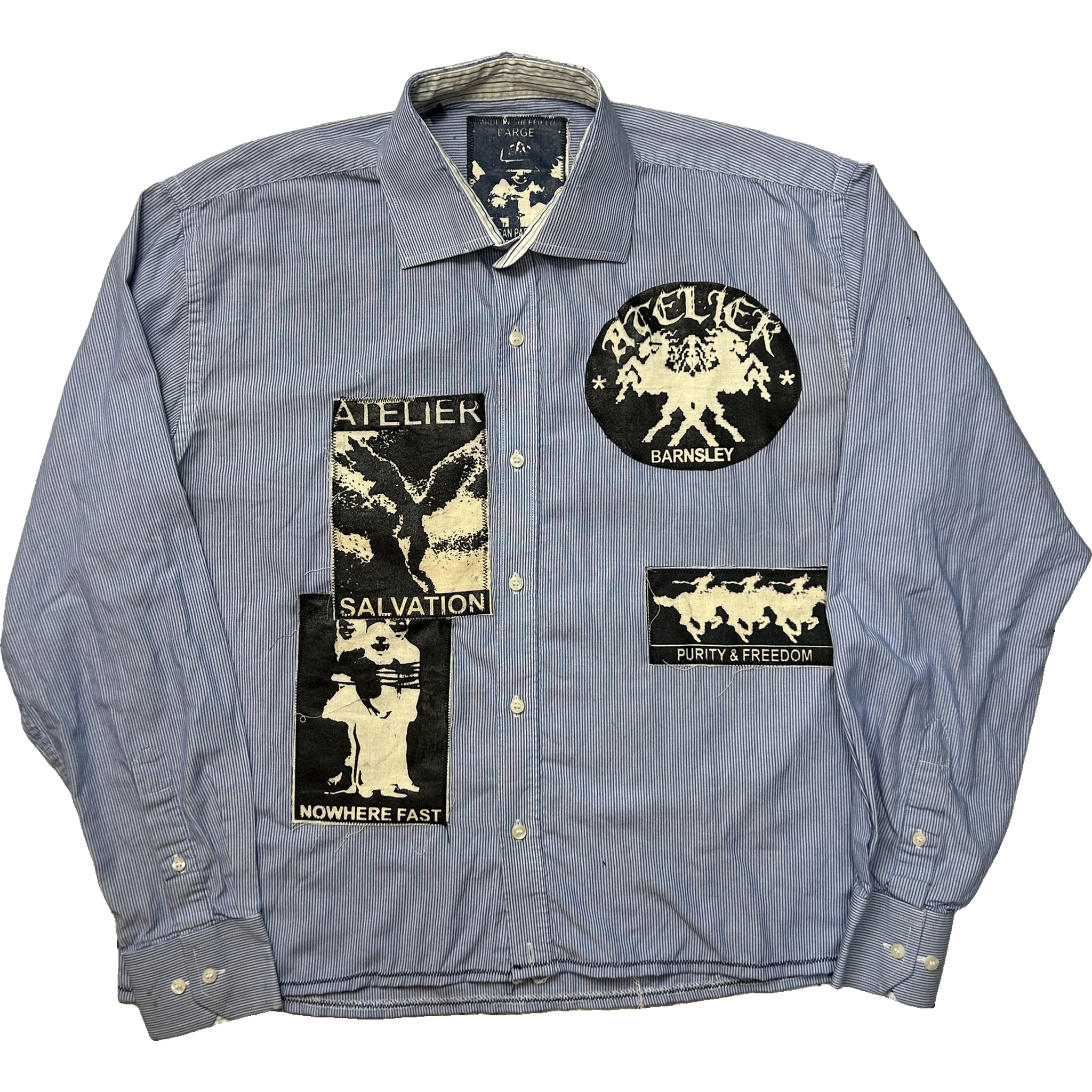 A light blue pinstriped button-up shirt with multiple black and cream graphic patches on the front, including a circular 'ATELIER NOWHERE FAST' patch and a 'PURITY & FREEDOM' patch.